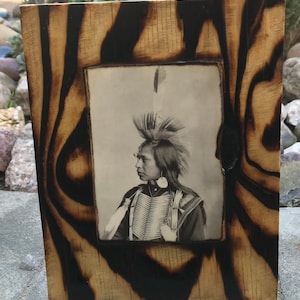May include: A black and white photograph of a Native American man in traditional clothing, framed in a wooden frame with a burnt wood finish.