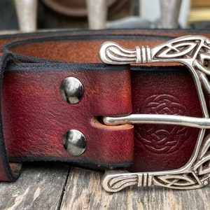 May include: A dark brown leather belt with a silver Celtic knot buckle and a silver Celtic knot design on the strap. The belt has a black stitching and silver rivets.