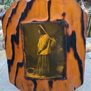 May include: A framed sepia-toned photograph of a person wearing traditional clothing and carrying a water jug. The frame is made of wood with a dark brown stain and a black border.