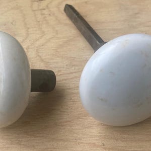 May include: Two white oval-shaped porcelain door knobs with dark metal stems. One knob is attached to a metal rod. The knobs are on a light brown wooden surface. The knobs are likely for a door or cabinet.