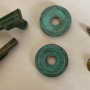 May include: An assortment of vintage door hardware components, including two handles, two circular escutcheons, a brass plate, and small screws. The hardware has a weathered, verdigris finish, suggesting age and use. The pieces are arranged on a light-colored surface.