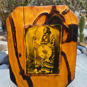 May include: A framed sepia-toned photograph of a Native American man in traditional clothing holding a shield and spear. The frame is made of wood with a dark brown stain.
