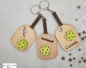 Op de afbeelding: Drie houten pickleball-racket sleutelhangers met een groen pickleball-balontwerp. De sleutelhangers zijn gepersonaliseerd met de namen "Pickleball", "Connie" en een bliksemontwerp.
