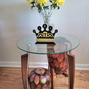 May include: A decorative table with a glass top, supporting a black and gold crown-shaped object. A vase of yellow flowers sits behind the crown. A round urn and a tall vase with a leaf pattern are also present.