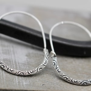 May include: A pair of silver hoop earrings with a decorative, floral design around the edge.