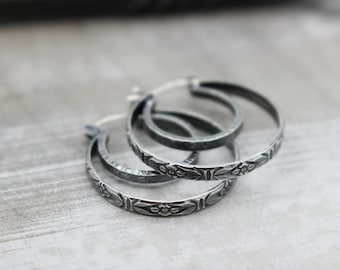 May include: Three silver hoop earrings with a floral design. The earrings are stacked on top of each other.