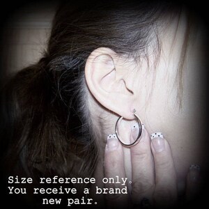 May include: A close-up of a person's ear with a silver hoop earring. The earring is about 1 inch in diameter. The person's hair is dark brown and pulled back in a ponytail.