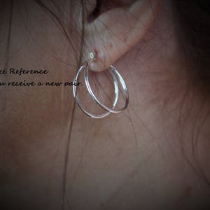 May include: Close-up of a person's ear wearing a pair of silver hoop earrings. The earrings are simple and elegant, with a smooth finish. The text "Size Reference You receive a new pair." is visible in the image.
