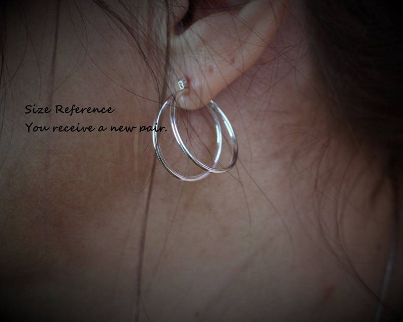 May include: Close-up of a person's ear wearing a pair of silver hoop earrings. The earrings are simple and elegant, with a smooth finish. The text "Size Reference You receive a new pair." is visible in the image.