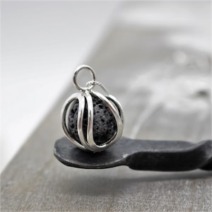 May include: A silver pendant featuring a black lava rock encased within a cage-like design. The pendant has a circular opening at the top.
