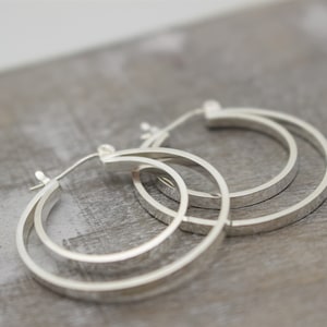 May include: A pair of silver hoop earrings with a double-layered design. The earrings are made of a smooth, polished metal and have a simple, elegant style.