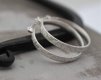 May include: A pair of silver hoop earrings with a delicate floral pattern. The earrings are made of sterling silver and have a polished finish.