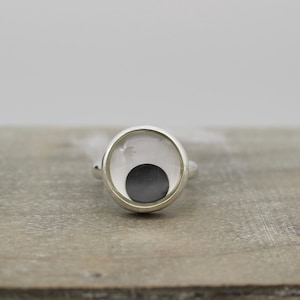 May include: A silver ring with a round, clear stone set in a silver bezel. The stone has a black circle in the center.
