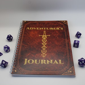 Könnte beinhalten: Ein Spiralbuch mit den Worten "ADVENTURER'S JOURNAL" in goldener Schrift auf einem braunen Einband. Der Einband zeigt ein goldenes Schwert und ein geometrisches Design. Sechs lila Würfel sind um das Journal verstreut.