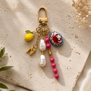 May include: Gold-tone keychain with a lobster clasp, featuring a yellow lemon charm, a gold letter "N", white pearl beads, pink beads, and a blue and white crab charm. The keychain is a decorative accessory.