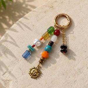 May include: A colorful beaded keychain with a gold-tone clasp. The keychain features a variety of beads in shades of blue, orange, green, and red, with a gold sun charm. The beads are strung together in a decorative pattern.