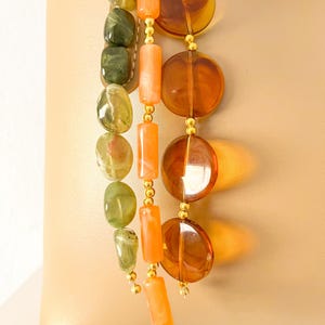 May include: A beige leather handbag adorned with three hanging beaded strands. One strand features olive green irregular beads, another has cylindrical orange beads, and the third consists of large, translucent amber-colored circular discs.