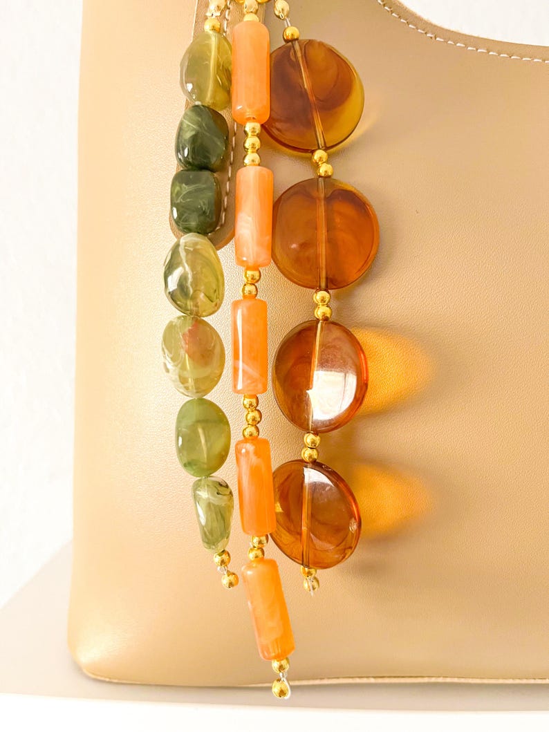 May include: A beige leather handbag adorned with three hanging beaded strands. One strand features olive green irregular beads, another has cylindrical orange beads, and the third consists of large, translucent amber-colored circular discs.