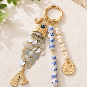 May include: Gold-toned keychain with a fish-shaped charm. The fish has iridescent scales and a gold tail. Additional charms include a seashell with a pearl, blue and white beads, and a gold coin. A stylish accessory.