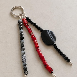 May include: A keychain featuring a silver-toned metal ring with three dangling strands. One strand has grey and black beads, another has irregular red coral-like segments, and the third has small black beads with a large, flat, oval black agate stone.