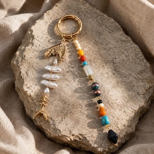 May include: A gold-colored keychain with a fishbone charm and a lobster claw charm. The keychain also features a strand of colorful beads, including white, red, yellow, blue, and black. The keychain is resting on a light brown rock.