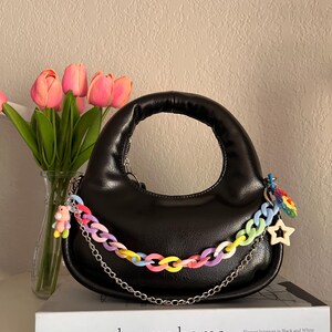 May include: Black handbag with a rounded shape and a short handle. A colorful chain strap and a silver chain are attached. The bag is decorated with a small bear charm and a star charm. The text "monochrome home" is visible on a book.