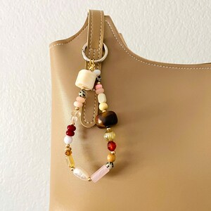 May include: A beaded bag charm with a variety of colorful beads, including cream, pink, brown, and clear beads. The charm is attached to a tan leather bag with a silver ring.