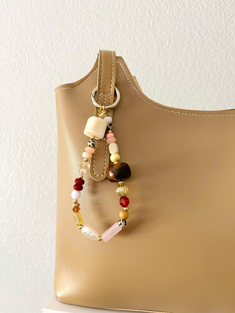 May include: A beaded bag charm with a variety of colorful beads, including cream, pink, brown, and clear beads. The charm is attached to a tan leather bag with a silver ring.
