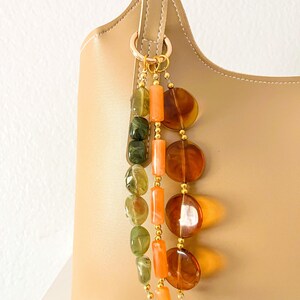 May include: A beige handbag adorned with a decorative charm featuring three hanging strands of beads. One strand has green marbled oval beads, another has orange cylindrical beads, and the third has large, translucent amber-colored circular discs.