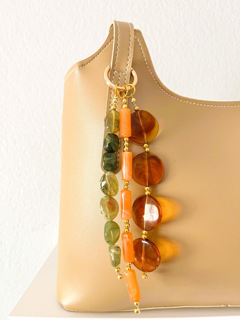 May include: A beige handbag adorned with a decorative charm featuring three hanging strands of beads. One strand has green marbled oval beads, another has orange cylindrical beads, and the third has large, translucent amber-colored circular discs.