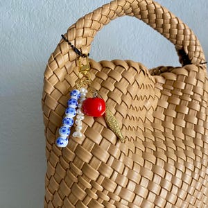 May include: A tan woven leather handbag with a braided handle. A gold-colored charm with a red apple, a fish, and blue and white beads hangs from the handle. The bag has a structured shape.