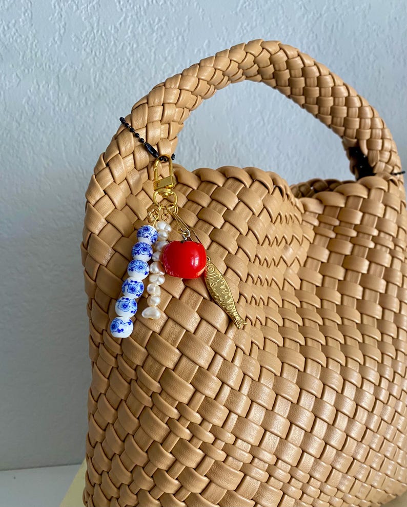 May include: A tan woven leather handbag with a braided handle. A gold-colored charm with a red apple, a fish, and blue and white beads hangs from the handle. The bag has a structured shape.