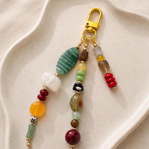 May include: A gold-toned metal lobster clasp keychain featuring three dangling strands of multi-colored beads. The beads include various shapes and sizes in shades of green, white, red, yellow, brown, and black, accented with small gold-toned metal spacers.