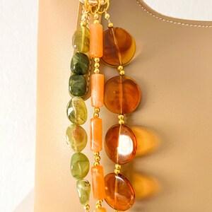 May include: A decorative bag charm featuring three hanging strands of beads attached to a gold-toned ring. One strand has green marbled oval beads, one has orange cylindrical beads, and one has large, translucent amber-colored circular discs.