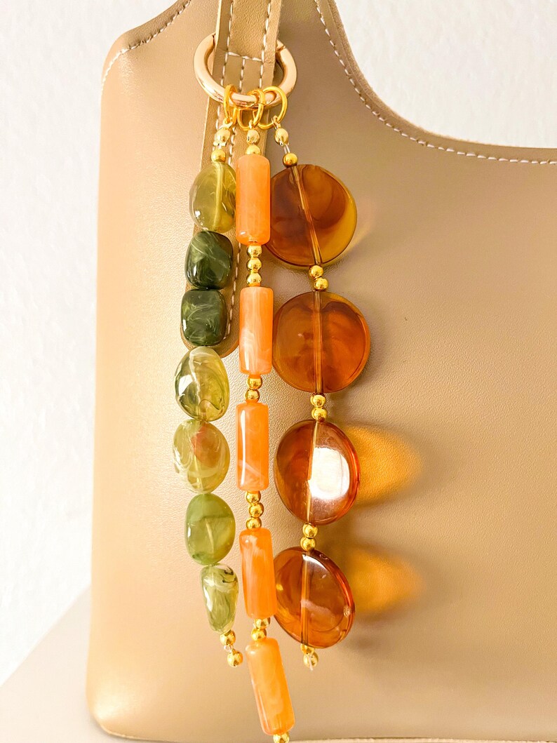 May include: A decorative bag charm featuring three hanging strands of beads attached to a gold-toned ring. One strand has green marbled oval beads, one has orange cylindrical beads, and one has large, translucent amber-colored circular discs.