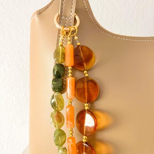 May include: A beige handbag adorned with a decorative charm featuring three hanging strands of beads. One strand has green marbled oval beads, another has orange cylindrical beads, and the third has large, translucent amber-colored circular discs.