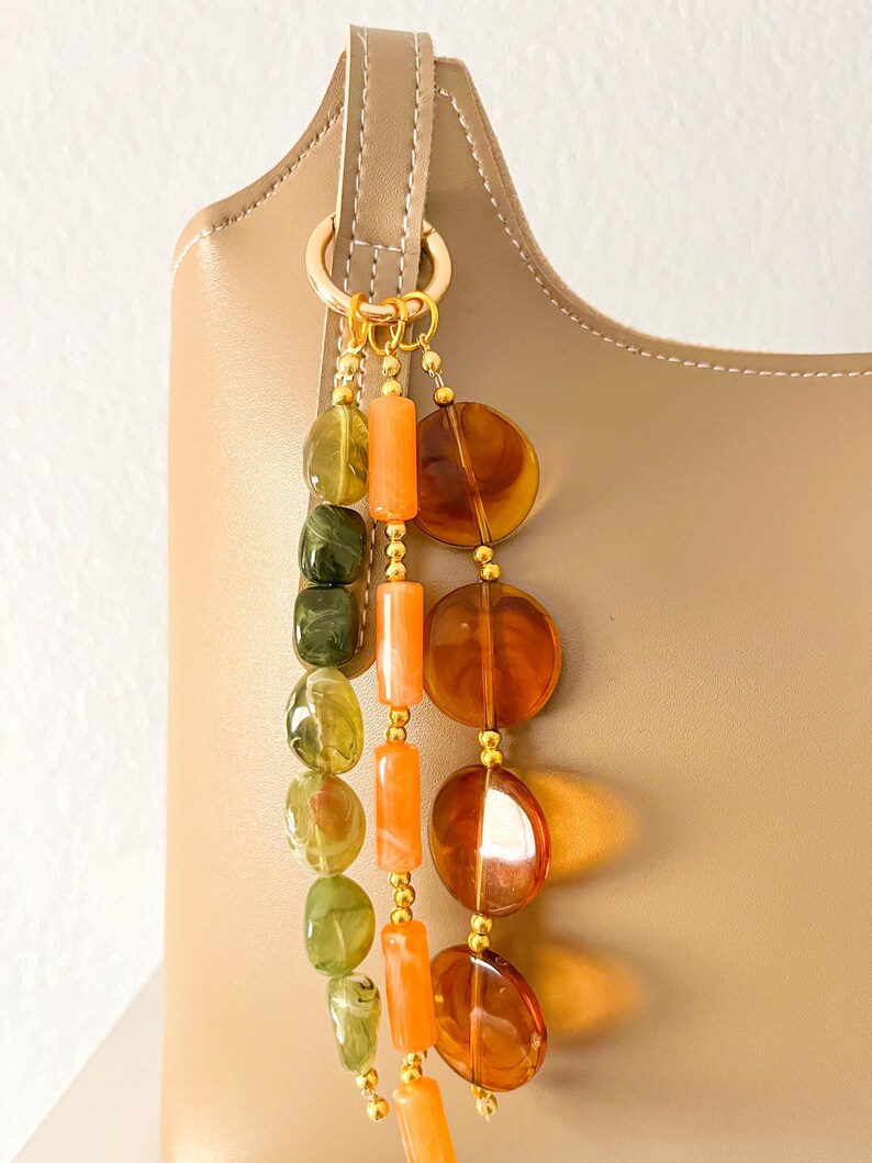 May include: A beige handbag adorned with a decorative charm featuring three hanging strands of beads. One strand has green marbled oval beads, another has orange cylindrical beads, and the third has large, translucent amber-colored circular discs.