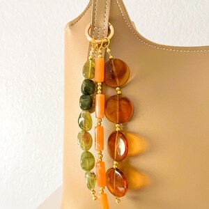 May include: A beige leather handbag adorned with a decorative charm featuring three hanging strands of beads. The strands include translucent green, opaque orange, and translucent amber-colored beads, all connected by gold-toned metal rings.
