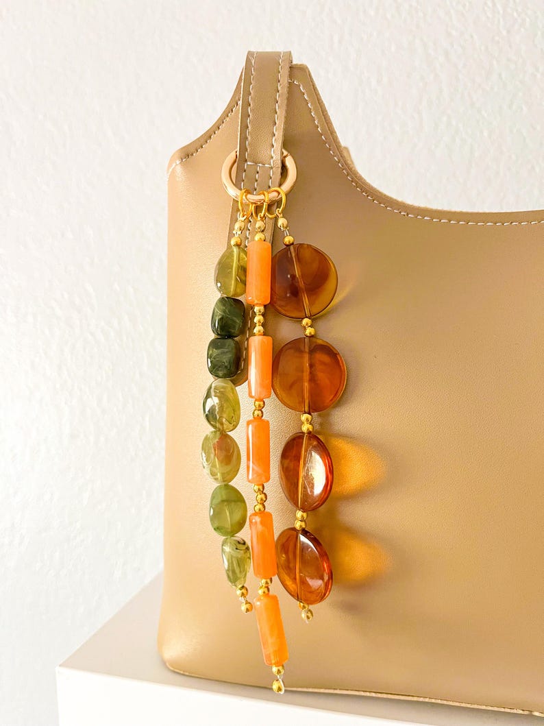 May include: A beige leather handbag adorned with a decorative charm featuring three hanging strands of beads. The strands include translucent green, opaque orange, and translucent amber-colored beads, all connected by gold-toned metal rings.