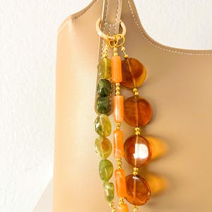May include: A beige handbag adorned with a decorative charm featuring three hanging strands of beads. The strands consist of green irregular stones, orange cylindrical beads, and large, translucent, circular amber-colored discs.