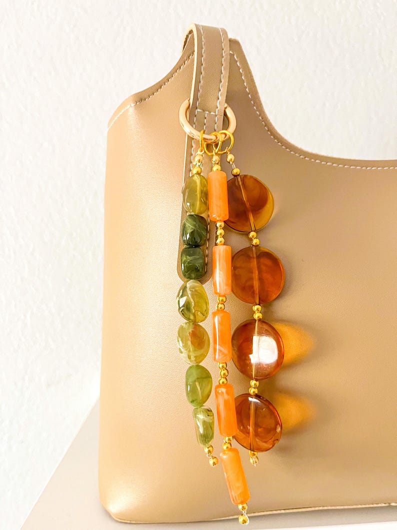 May include: A beige handbag adorned with a decorative charm featuring three hanging strands of beads. The strands consist of green irregular stones, orange cylindrical beads, and large, translucent, circular amber-colored discs.