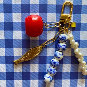 May include: A gold-toned keychain with a red cherry charm, a gold fish charm, and a strand of white and blue floral beads. The keychain also features a small yellow heart charm and a strand of white pearls.