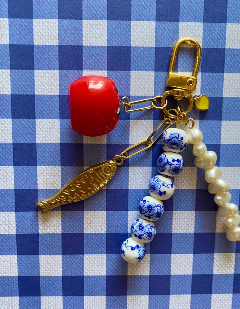 May include: A gold-toned keychain with a red cherry charm, a gold fish charm, and a strand of white and blue floral beads. The keychain also features a small yellow heart charm and a strand of white pearls.