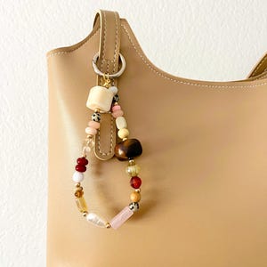 May include: A beige handbag with a beaded charm. The charm features a variety of colorful beads, including pink, brown, white, and gold, attached to a metal ring. The bag has a simple, structured design.