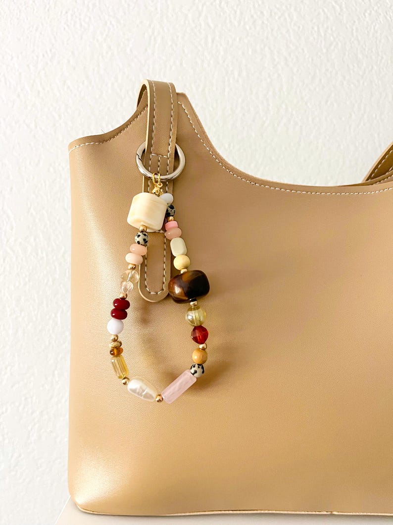 May include: A beige handbag with a beaded charm. The charm features a variety of colorful beads, including pink, brown, white, and gold, attached to a metal ring. The bag has a simple, structured design.