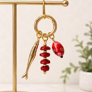 May include: Gold-toned keychain with three charms: a fish, a stack of red beads, and a red oval bead with black accents. The keychain is attached to a gold-colored metal stand, with a plant in the background.