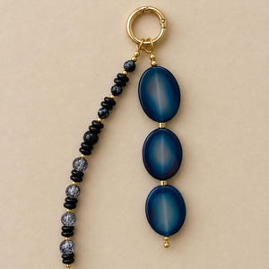 May include: A gold-toned metal keyring featuring two dangling strands. One strand consists of small black and grey speckled beads, while the other features three large, smooth, oval-shaped blue beads separated by small gold-toned metal spacers.
