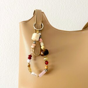 May include: A tan leather handbag with a beaded charm. The charm features a variety of beads in colors like pink, red, yellow, and brown. The beads are strung together on a loop and attached to the bag's handle.