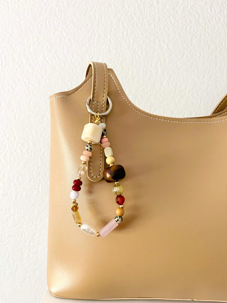 May include: A tan leather handbag with a beaded charm. The charm features a variety of beads in colors like pink, red, yellow, and brown. The beads are strung together on a loop and attached to the bag's handle.