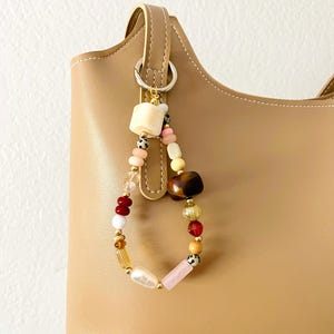 May include: A beaded keychain with a variety of colorful beads, including white, pink, red, and gold, hangs from a tan handbag. The beads are various shapes and sizes, creating a unique and eye-catching accessory.